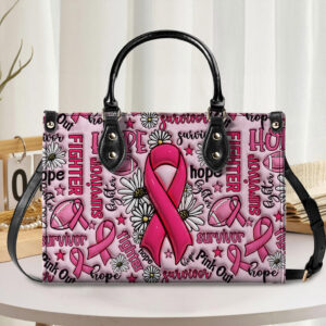 Breast cancer handbag in pink with black handles, featuring floral and ribbon designs, front view on a table.