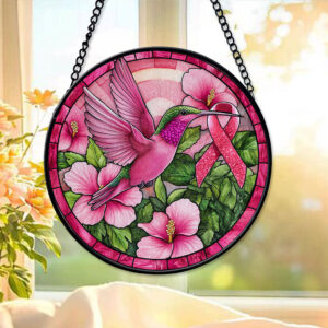 Front view of a faux stained glass pink ribbon suncatcher showing a pink hummingbird and flowers hanging in a bright window.